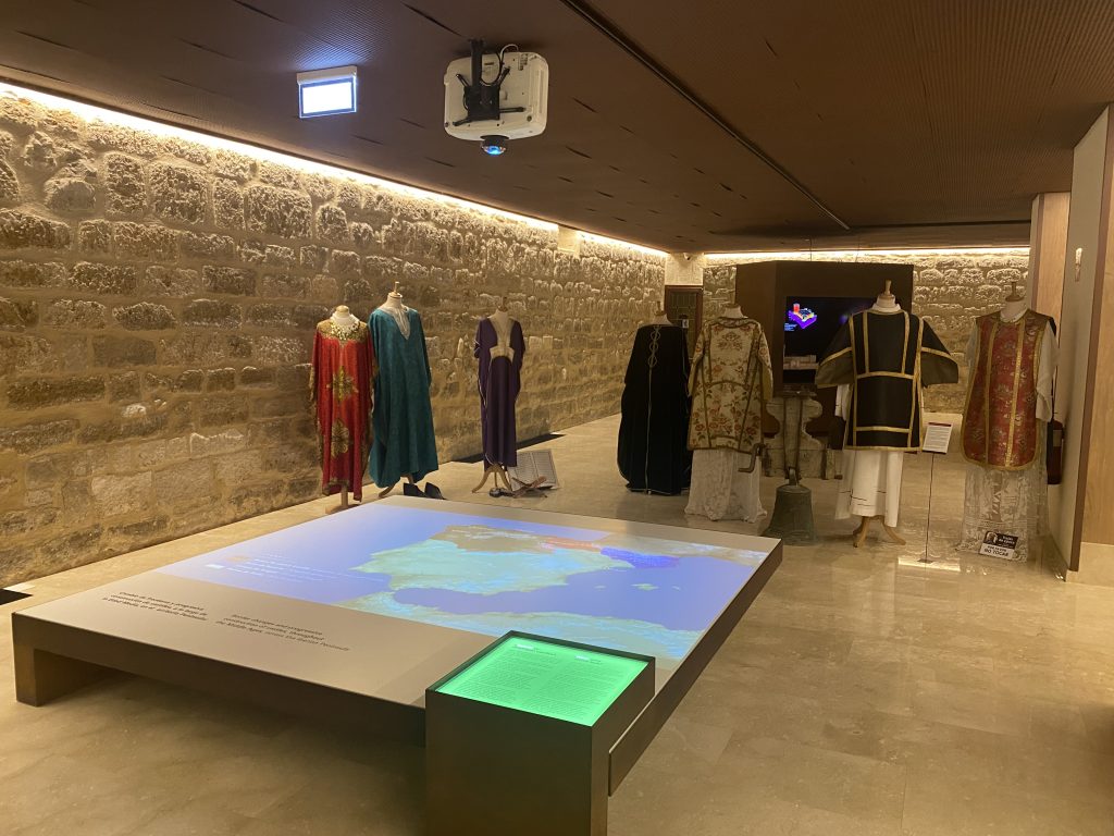 Sala del museo con pantalla interactiva situada en el suelo en la que se ve un mapa de España. Al fondo se ven diferentes trajes de época de hombre y de mujer. 