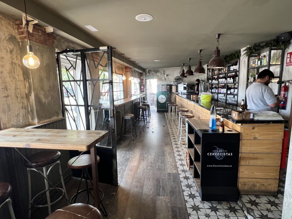 Interior de restaurante, a lado derecho se ve la barra del bar y al lado izquierdo mesas altas con taburetes