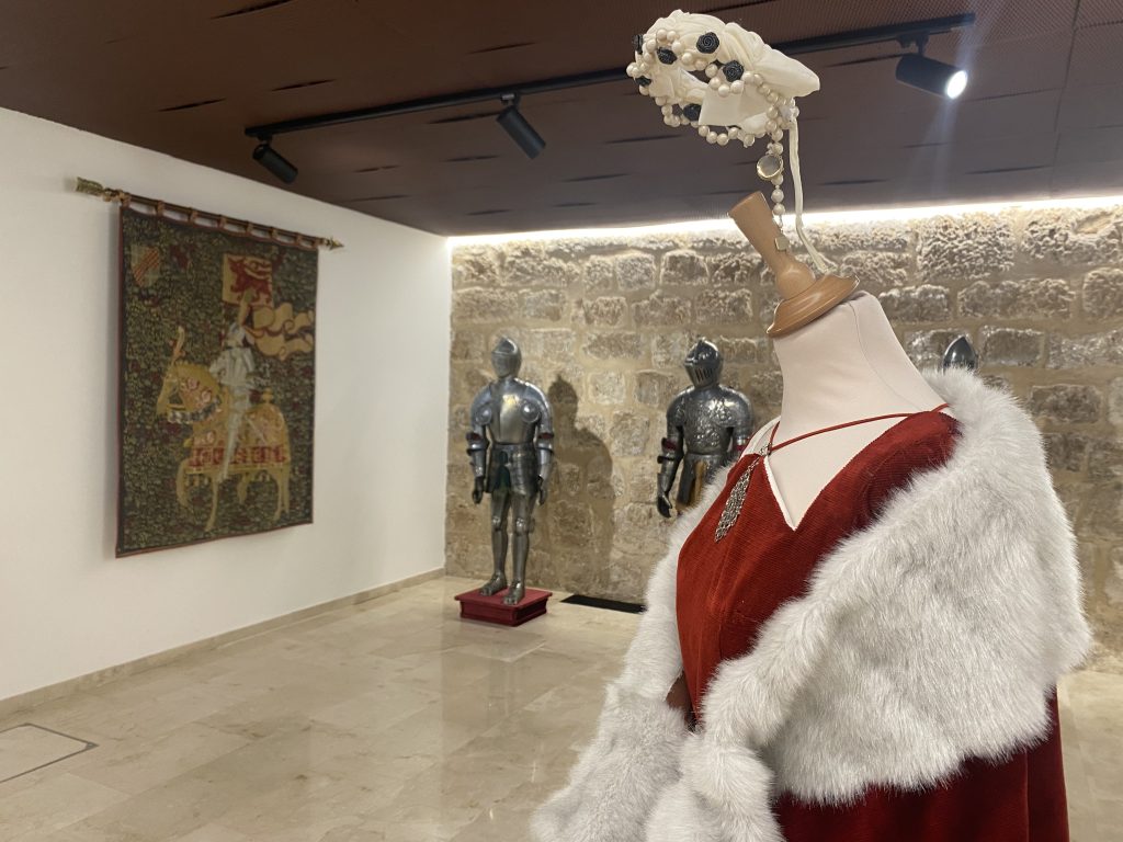 Sala del museo con armaduras de caballeros, tapiz colgado en la pared y vestido de época de mujer. 