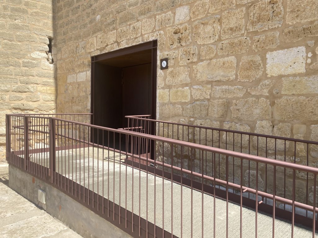 Acceso de entrada al castillo con rampa de cemento con pasamanos de hierro y puerta de doble hoja de más de 80 cm. 