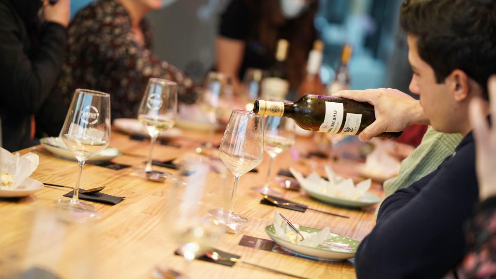 Mesa de sala de catas con personas haciendo una cata donde se ven copas, platos y una botella de vino echando vino en una copa. 