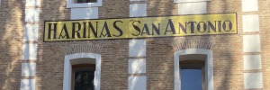 La imagen es el letrero de la fábrica, todo mayúscula Harinas San Antonio. Letras negras sobre fondo amarillo. El letrero está colocado sobre un edificio de ladrillos.