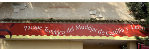 Es el letrero del Parque Temático del Mudéjar de Castilla y León. El fondo es de dos franjas de colores separados por una línea ondulada. La franja superior es de color rojo y es más ancha. La inferior es más estrecha, de color amarillo.